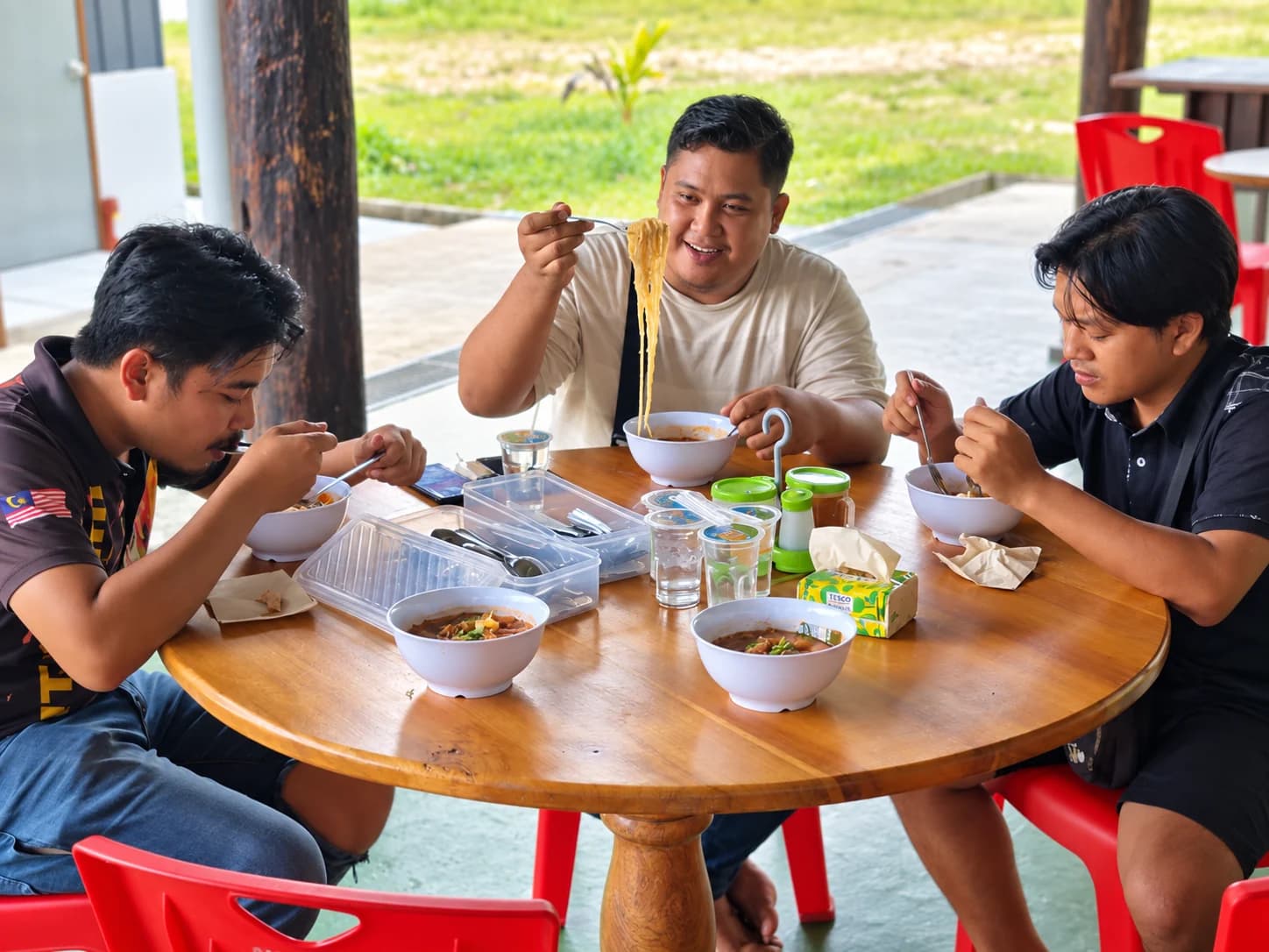 A diner at D’Cafe grinning as he lifts a forkful of noodles high above his bowl, with two others eating noodles beside him