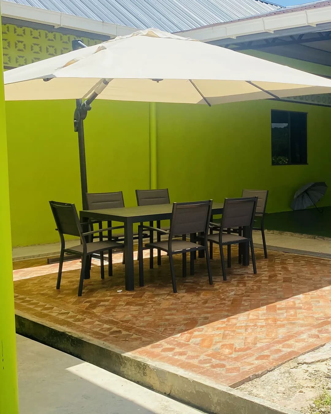 A big cream umbrella over a table and chairs on the brick patio beside D’Cafe, with the covered dining area and its red chairs behind