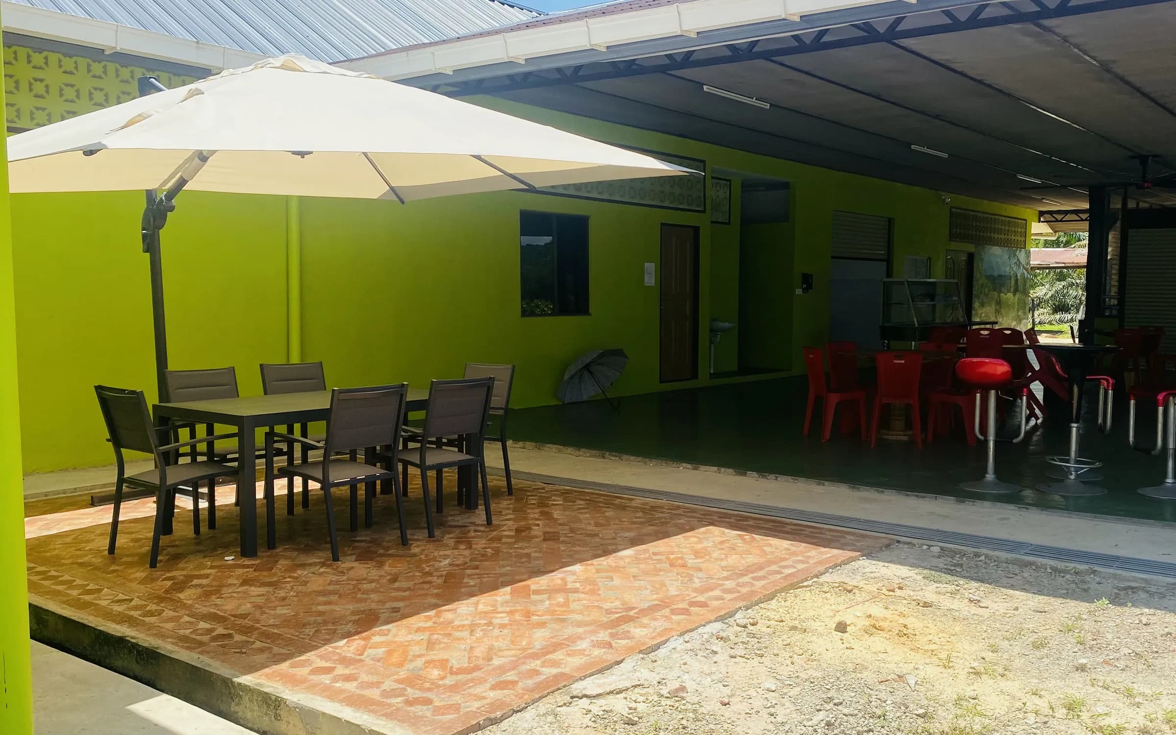 A big cream umbrella over a table and chairs on the brick patio beside D’Cafe, with the covered dining area and its red chairs behind